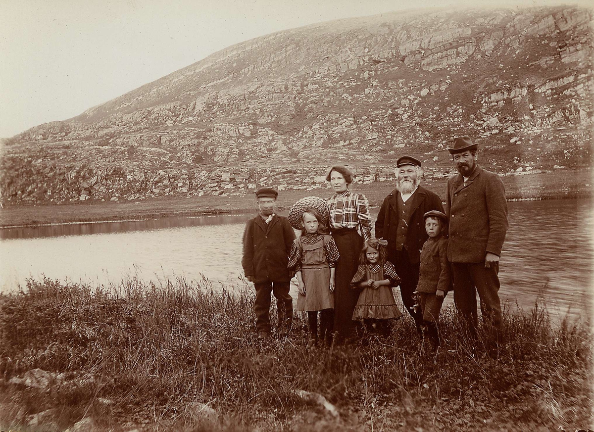 Persongruppe stående i fjordlandskap, Båtsfjord, 4 voksne og 3 barn der kvinnen er Hanna Resvoll-Holmsen selv. 
Del av serie fra en forskningsreise i Øst-Finnmark 1909.