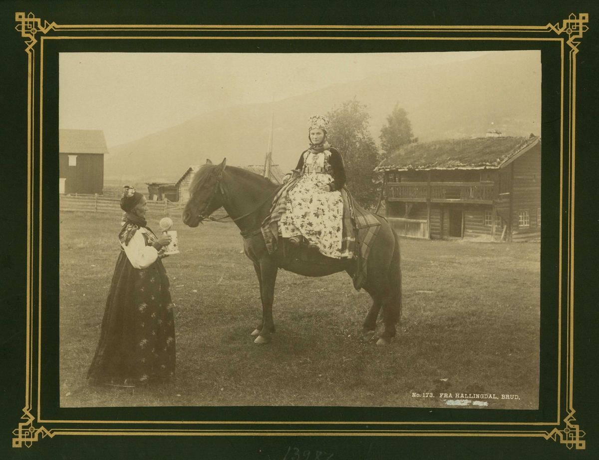 Kvinnedrakt, brud fra Hallingdal til hest på gårdstun, Buskerud.  Kvinne står ved siden av med ølkrus i hendene.
