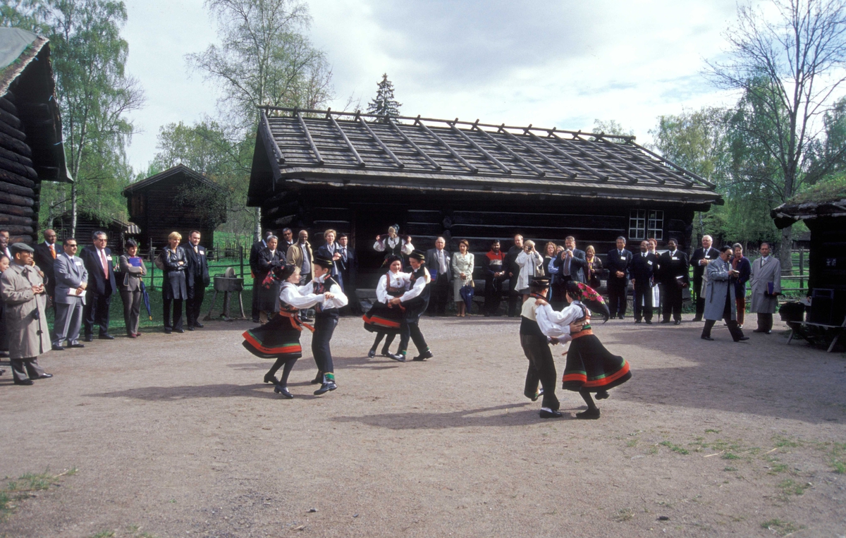 Norsk Folkemuseums dansegruppe, kledd i  setesdalsdrakter, danser folkedans i Numedalstunet på Norsk Folkemuseum. Publikum står i bakgrunnen og ser på.