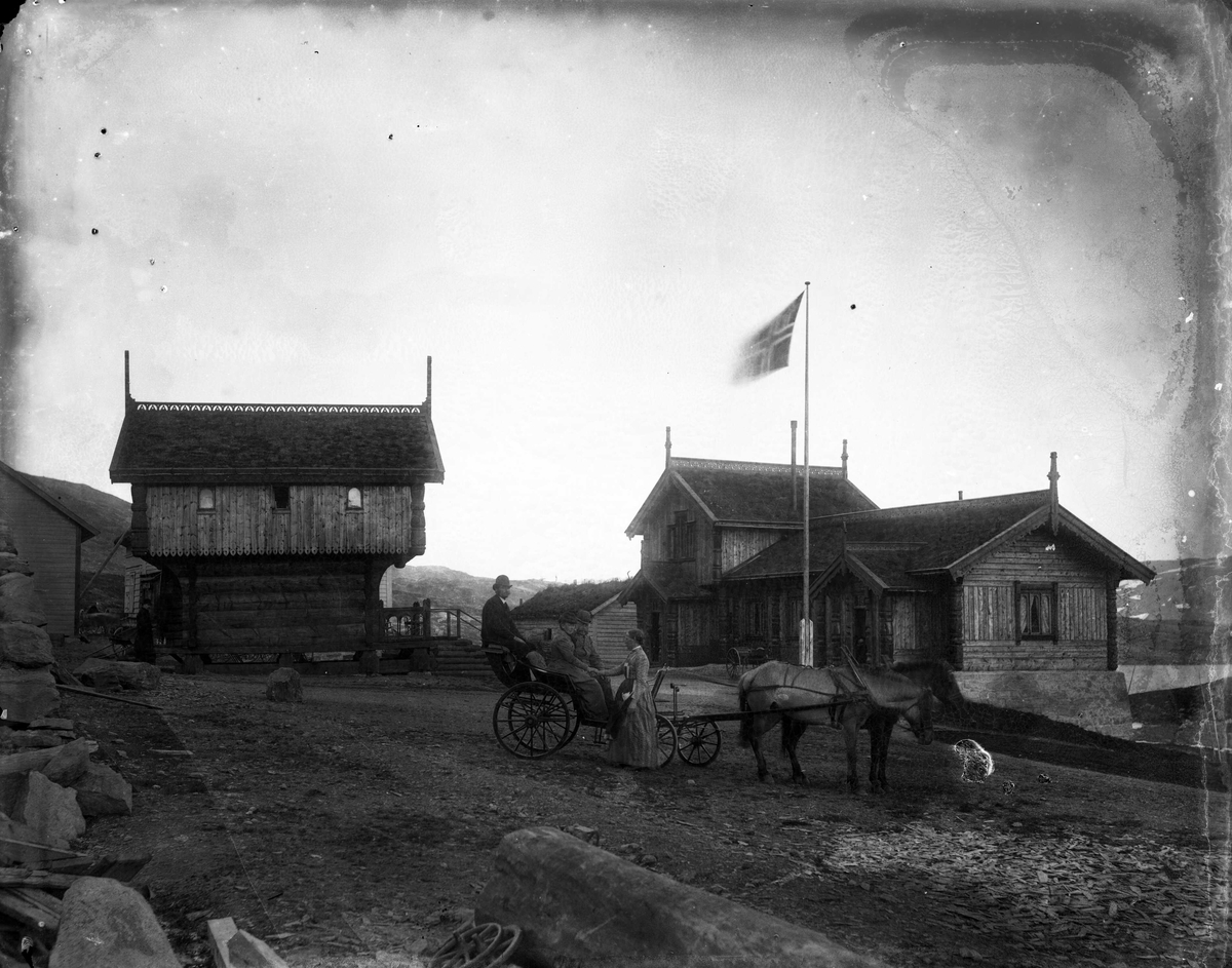 Gårdstun på Haukeliseter i Telemark, med personer sittende i en vogn trukket av to hester. Bygning, stabbur og flagg i bakgrunnen.
