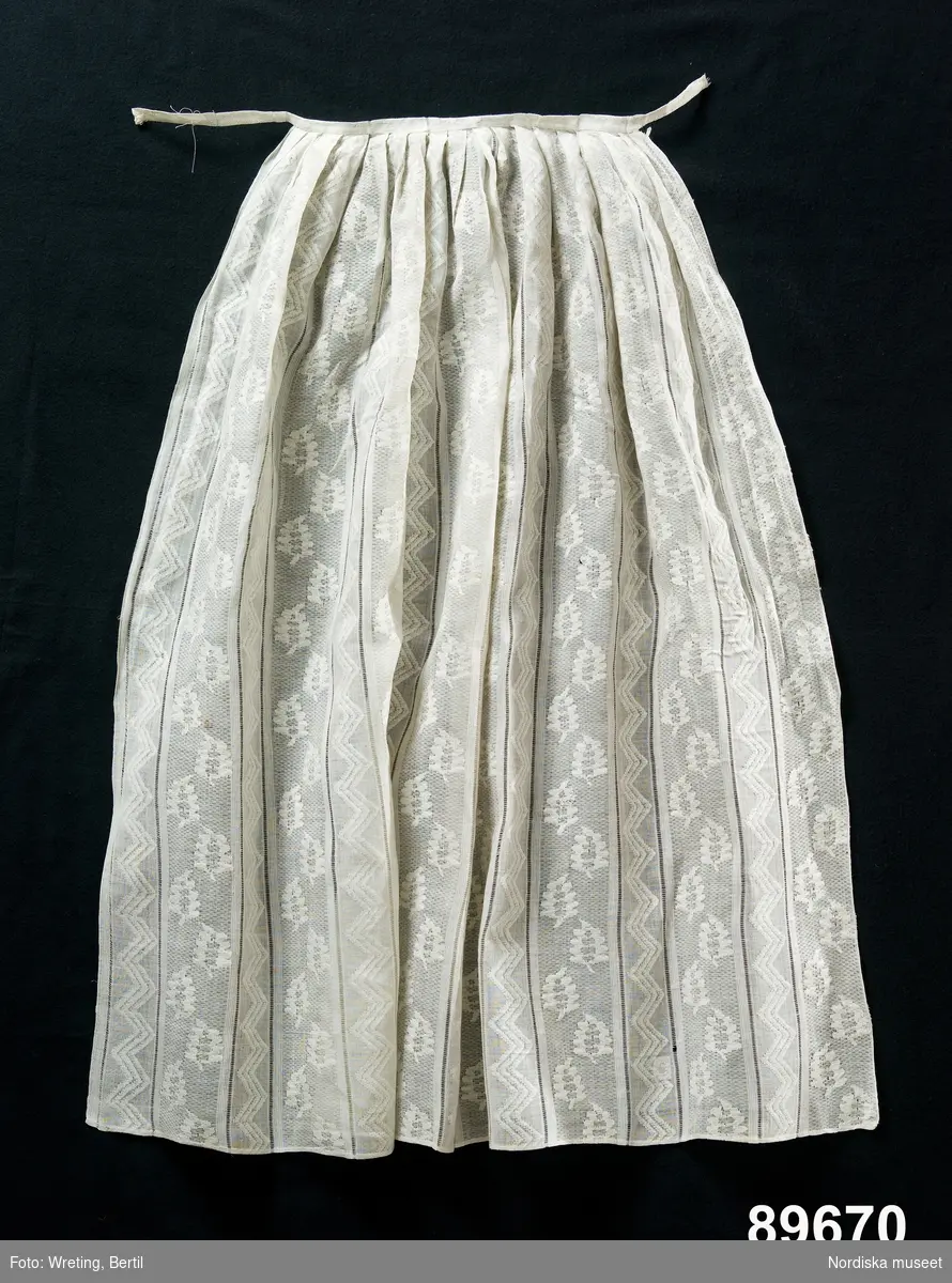 Förkläde, enligt uppgift brudförkläde från 1830-talet, vitt, maskinvävt bomullstyg i ränder med omväxlande sicksackband och blad, upptill veckat och försett med midjeband.

/Berit Eldvik maj 2006
