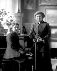 Gruppeportrett, to kvinner i frelsesarmeuniform. En ved pian