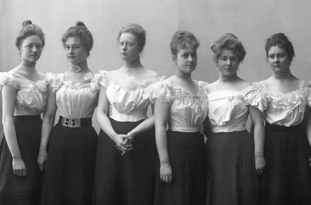 Gruppeportrett, syv kvinner i hvite kortermede bluser og svart skjørt.