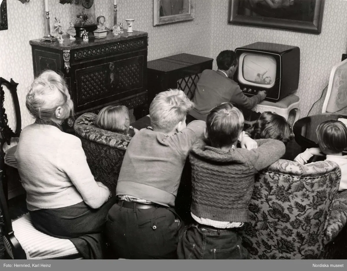 Barn och vuxna sitter och tittar på TV i vardagsrum.