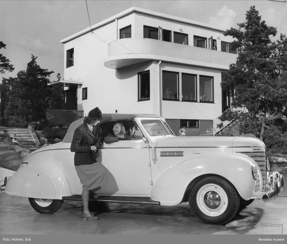 Kvinna i samtal med en passagerare i en vit Plymouth 1939, cabriolet, parkerad framför ett hus i funktionalsitisk stil. Nordiska Kompaniet.
