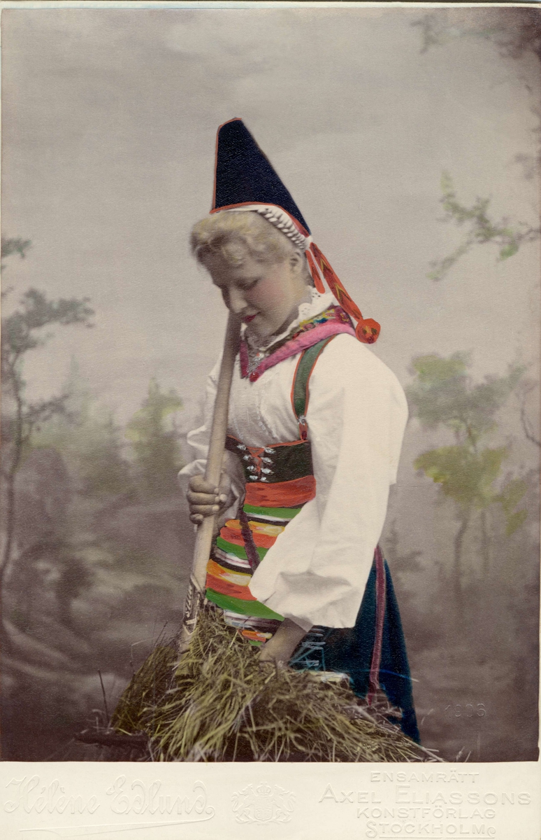 En kvinna klädd i folkdräkt från Dalarna , står med en räfsa och en hötapp. Handkolorerat ateljéporträtt. Klädd i Rättviksdräkt, som den visades i nationalromantisk framställning.