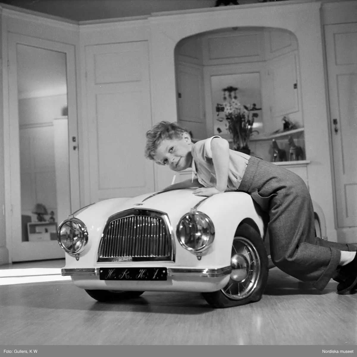 Kronprins Carl Gustaf leker med en trampbil. På nummerplåten 