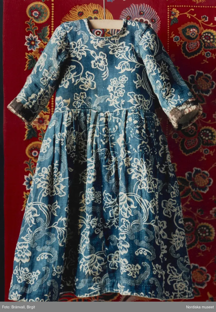 Barnkolt i blåtryck, sekelskiftet 1700-1800, i bakrunden rödbottnad kattun från schweiziskt tygtryckeri. 
Nordiska museets föremål inv.nr 3206.