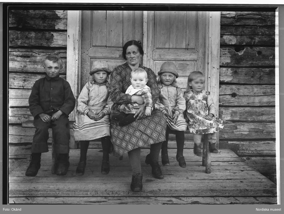 Kvinna med fem barn. Porträttbild av Amanda Ek född 1893 med sonen Sune född 1929 i famnen omgiven av döttrarna Signe (född 1924 tv i bild) och Henny född 1925, längst till vänster sonen Östen, som börjat skolan. Flickan längst till höger är en grannflicka. Gruppen sitter på en bänk på förstubron till ett timrat hus med dubbeldörrar i bakgrunden.