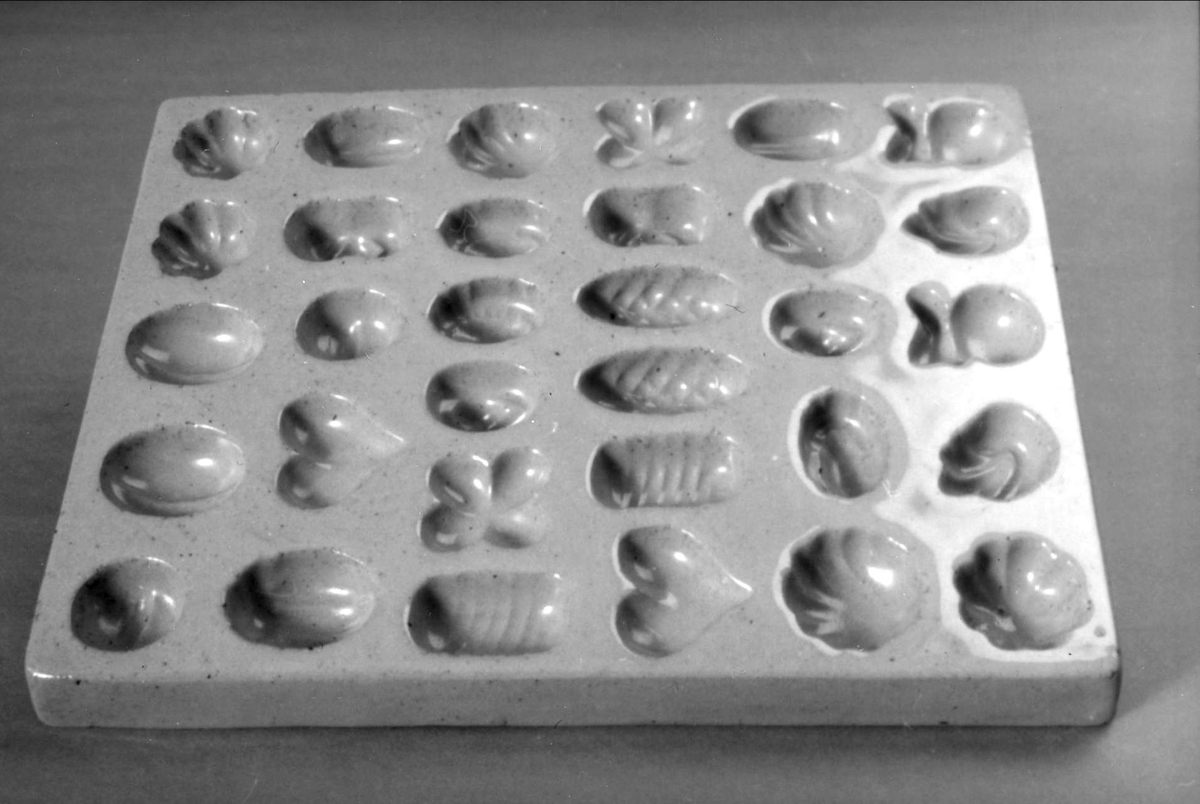 Fyrkantig form av gulglaserat lergods avsedd för tillverkning av marsipankonfekt med plats för 30 konfektbitar i form av 13 olika frukter. Stämpel på undersidan: S:T ERIKS LERVARUFABRIKER UPSALA. 
Denna stämpel användes före 1925.
