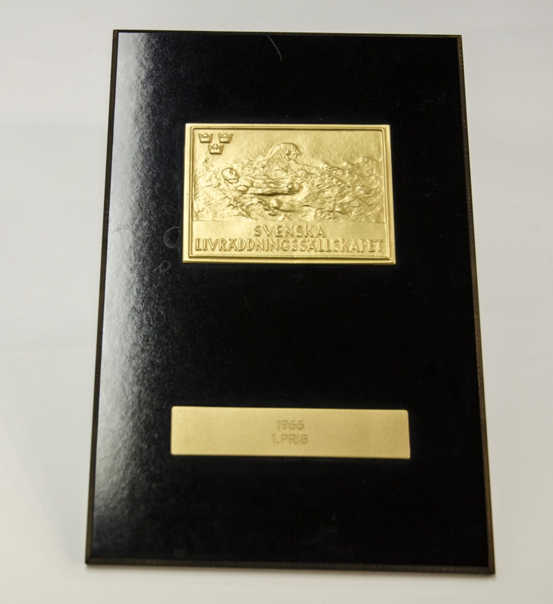 Guldfärgad metallplakett föreställande simmande man i lågrelief, i översta vänstra hörnet tre kronor samt texten: "SVENSKA LIVRÄDDNINGSSÄLLSKAPET", fästad på svart träplatta. Graverad med följande text: "1966 1. PRIS".