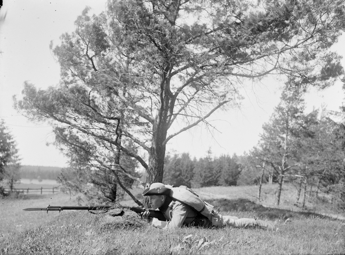En armésoldat lägger an ett gevär liggandes på en kulle. Mannen är Henning Axfors och tillhörde antagligen I 4 eller I 5 regemente.