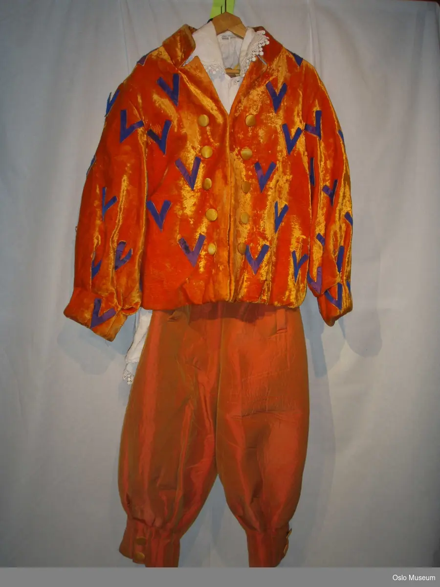 A=  jakke i oransje silkestoff med lilla V-formet dekor 
B= bukse i oransje boblestoff med gule knapper og bukseseler.
C= bluse i hvitt lin med kniplinger og stoffknapper