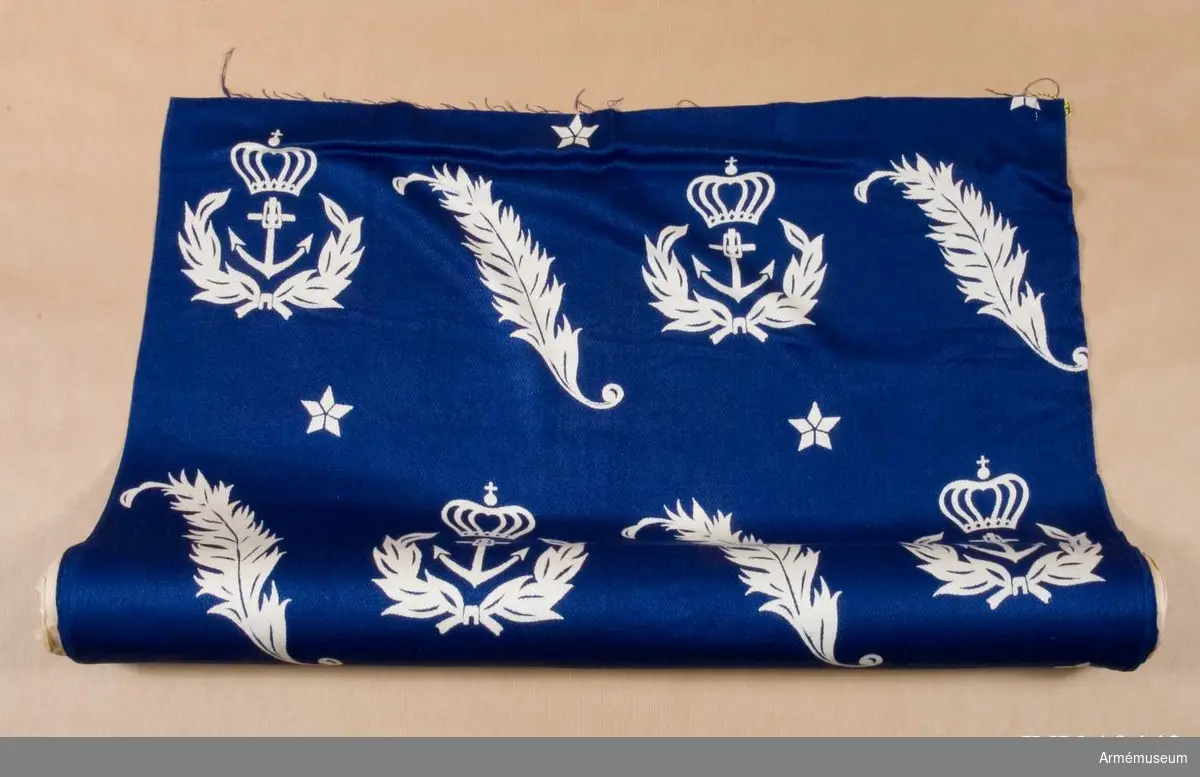 Tygprov från 1940-talet. Tillverkat för marinstaben. Formgivet av Astrid Sampe.
Textiltryck i vitt på en blå botten med motiv av ett ankare i mitten med kvistar runt om och krönt av kunglig krona. Det andra motivet är en yvig kvist och däremellan femuddiga stjärnor.

Samhörande gåva: 19441-2, tygprover.