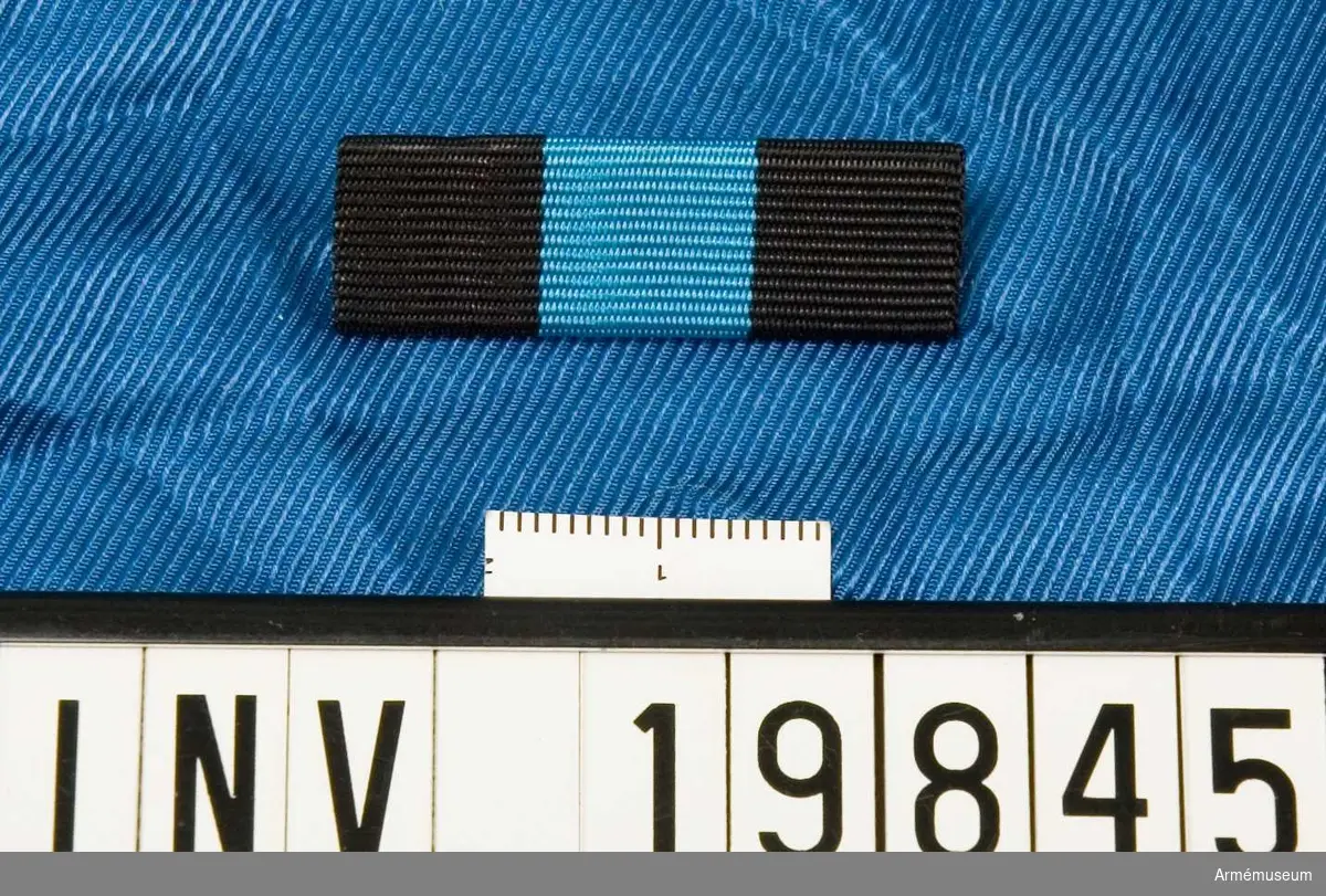 Band kluvet i svart, blått och svart. Släpspännet förvaras i ett etui tillsammans med en medalj och en miniatyrmedalj.