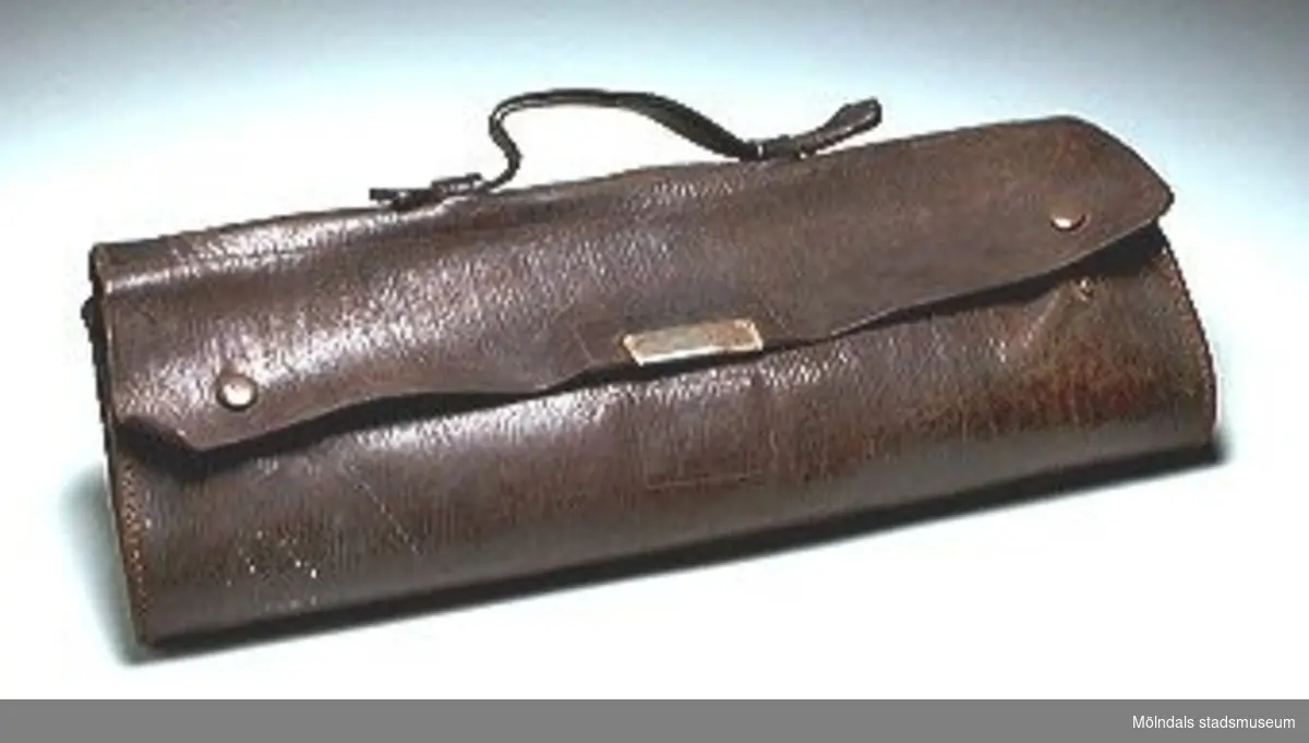 Mörkbrun portfölj med övervikt lock som knäpps i metallspänne på framsidan, handtag och tryckknappar.