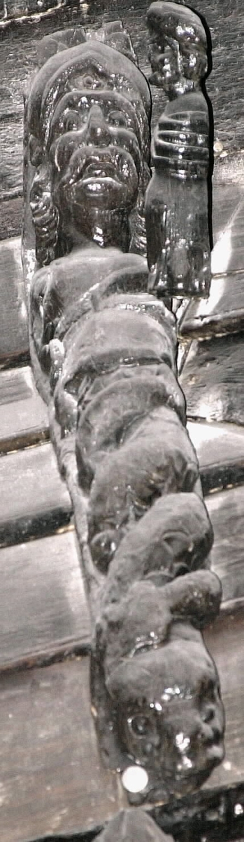 Triton med stora mustascher och lockigt hår. På huvudet sitter en hjälm med tudelad hjälmkam och nedhängande halsskydd i form av överlappande metallplåtar. Den rustningslika klädedräkten har en kvadratisk urringning. Kring midjan sitter ett band eller bälte. Den högra sidan av skulpturens bål upptas av en bladslinga som ersätter höger arm samt en del av midjebandet. Skulpturens fiskstjärt slingrar sig kring stjärten till en fisk eller en delfin vars huvud är synligt vid skulpturens fotända.

Skulpturens vänstra sida är inte skulpterad utan har utgjort en slät anliggningsyta som sträcker sig ned till det att fiskstjärten tar vid. Anliggningsytan är uppdelad på ett högre parti samt ett lägre part. Partierna är avgränsade genom ett vinkelformat hak i höjd med midjan. I det övre partiet återfinns en kvarsittande dymling. Bakstycket är kraftigt och snidat i ett med skulpturen. Skulpturens profil är svagt S-formad.
Skulpturen är relativt välbevarad.

Text in English: Triton with large moustache and long, curly hair. On the head he wears a smooth-surfaced helmet with upturned brim and hanging neck protector. A rudimentary crest can be seen on top of the helmet. The armour-like outfit has a square neck. There is a rudimentary girdle or belt round the waist. The arms are replaced by leafy ornamentation. Fastening surface opposite side left.

The fish tail is entwined with the tail of a fish or a dolphin which can be seen at the sculpture''s foot end. The figure is carved in one piece with a robust backboard. The profile is slightly S-shaped.
The sculpture is rather well preserved.