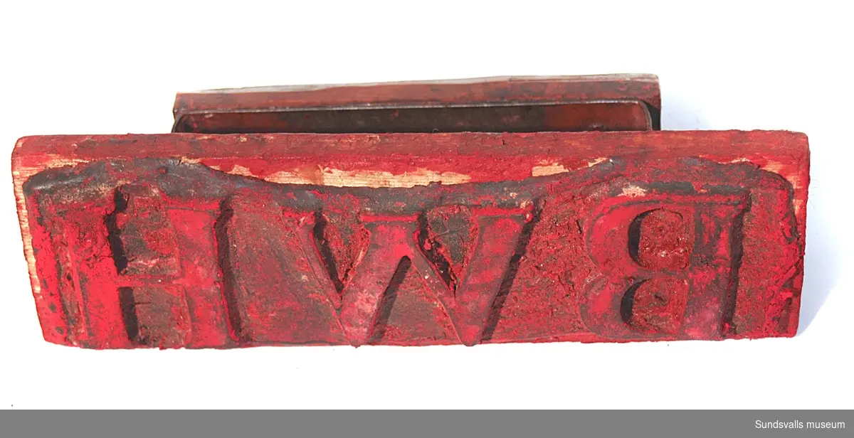 Stämpel i gummi med handtag i trä. Stämpeln visar bokstäverna BWH. Röda färgrester finns kvar på stämpeln.