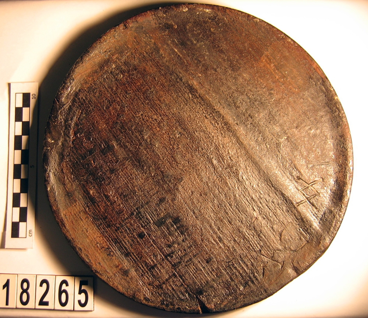 En svarvad trädisk. Disken har fem bomärken, två sitter på diskens ena sida, ett på andra sidan samt två stycken längs kanten.