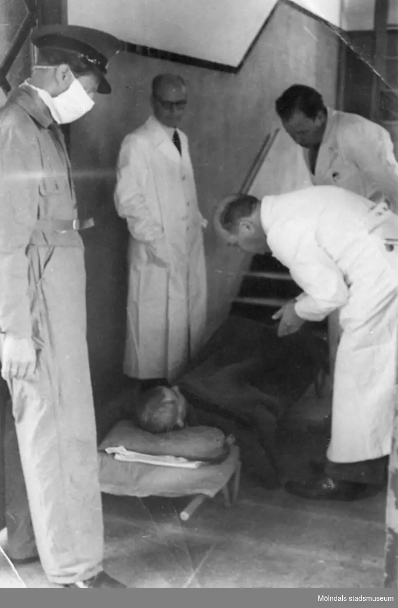 "Karantänsförläggning" på Kvarnbyskolan för överlevande från koncentrationsläger i Tyskland och Polen 1945.  
Skolan fungerade under denna sommar som beredsskapssjukhus.

En överlevande ligger på en bår, omgiven av tre läkare och en Röda Kors-arbetare.