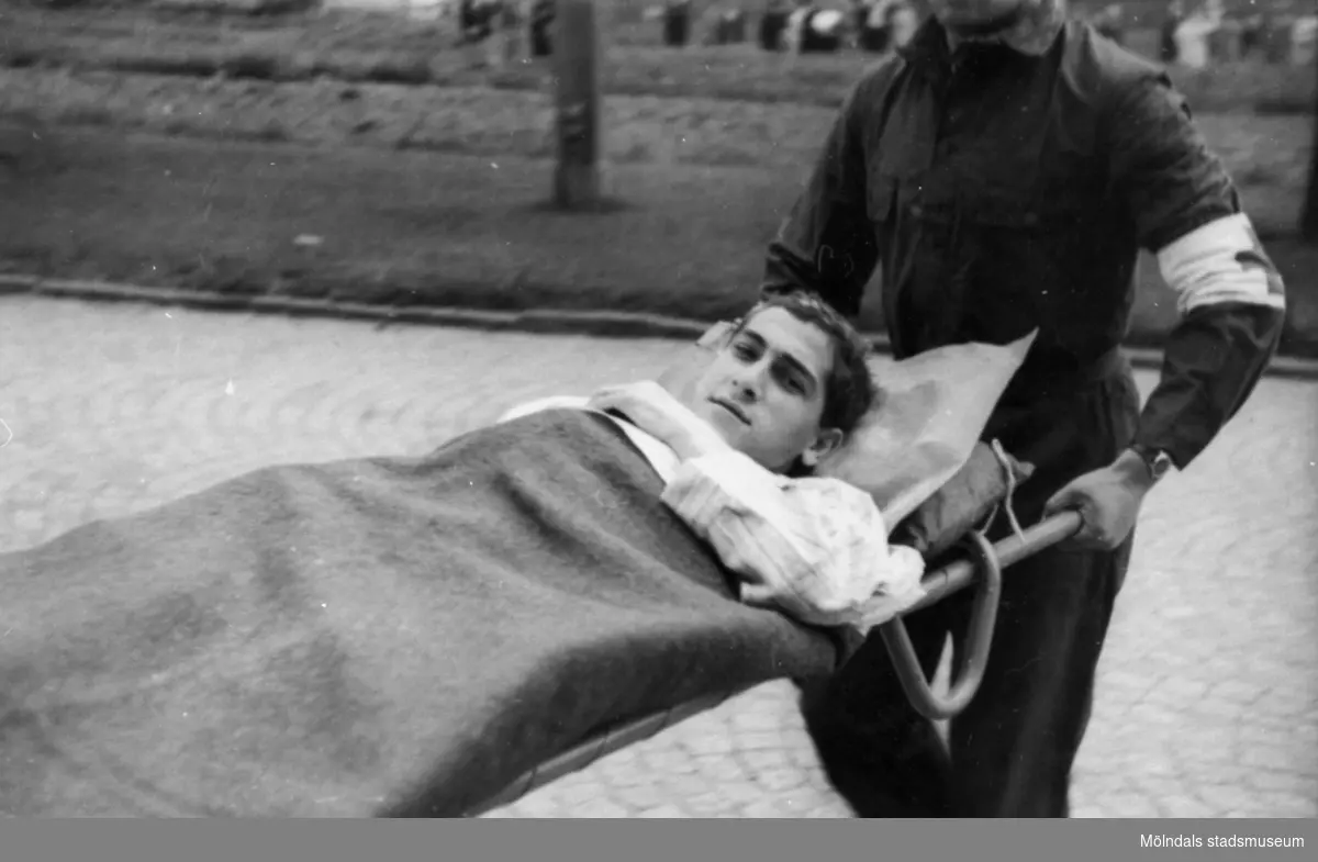 "Karantänsförläggning" på Kvarnbyskolan för överlevande från koncentrationsläger i Tyskland och Polen 1945.  
Skolan fungerade under denna sommar som beredsskapssjukhus.

En ung man blir buren på bår av Röda Kors-personal.