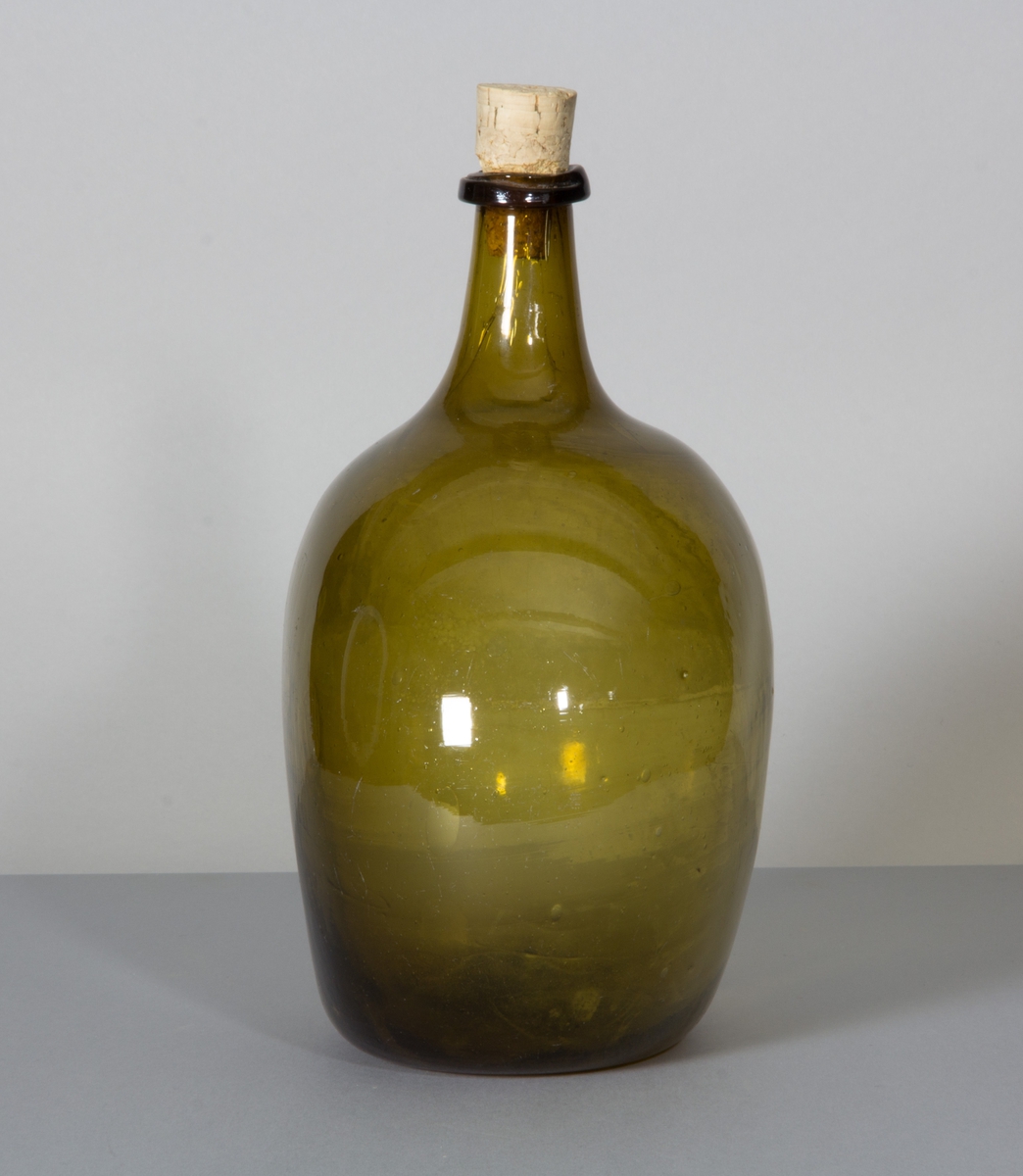 Flaska i glas, brungrön till färgen. Rundade sidor, hals med halsring och platt botten.