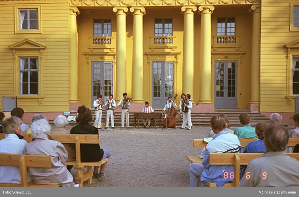 En orkester, bestående av sju män, spelar musik inför publik på Gunnebo slotts framsida. Publiken sitter på bänkar.