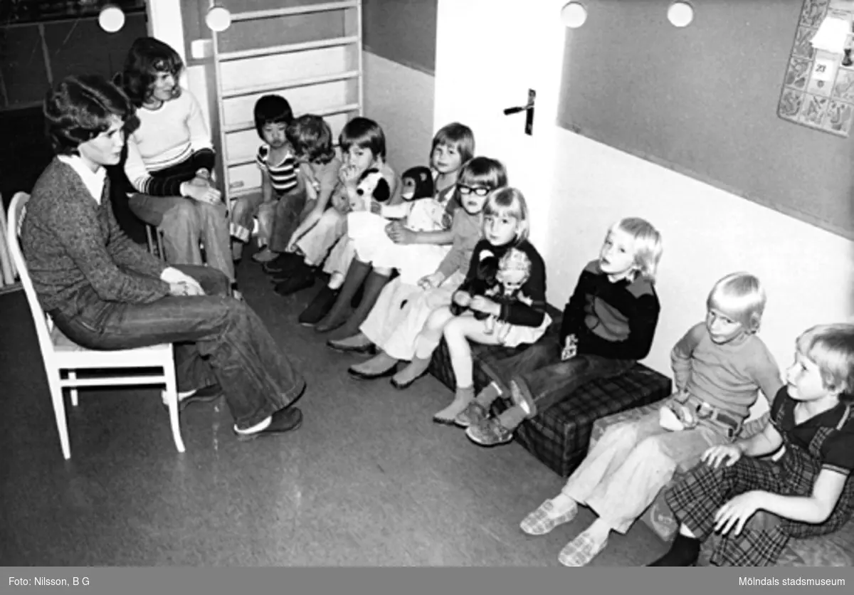 Barn som sitter på en bänk vid väggen och två fröknar som sitter på var sin stol mittemot barnen. Holtermanska daghemmet maj 1975.