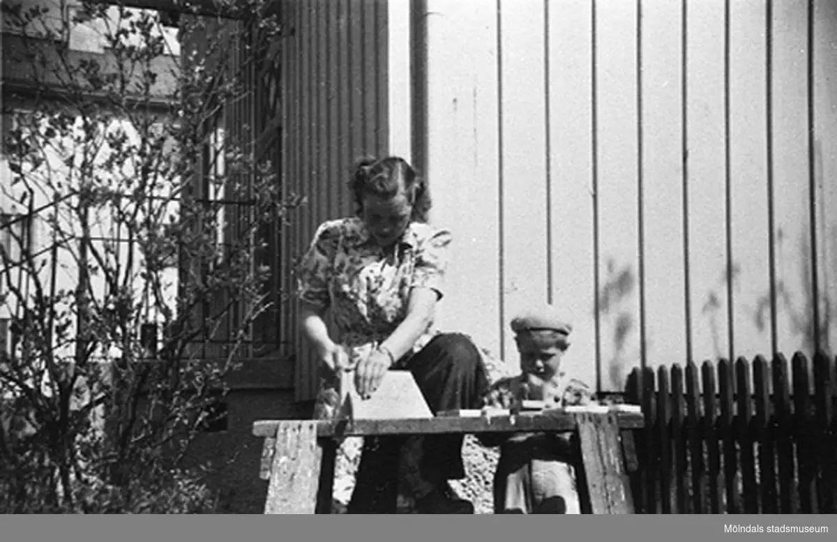 En lärare och ett barn sågar någonting i trädgården vid

Krokslätts daghem, Dalhemsgatan 7 i Krokslätt år 1949.