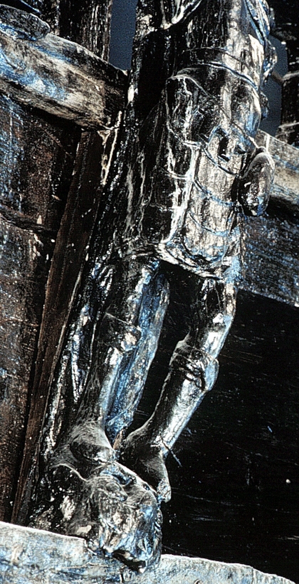 Skulptur föreställande en mansfigur iförd thoraxrustning, med en mantel draperad över bröstet. Manteln är fästad på höger axel med ett spänne. Figurens ansikte är slätrakat och på huvudet sitter en lagerkrans.
Figurens huvud är vridet något åt vänster. Bål och ben är riktade framåt. Vänster hand, som är snidad ur samma stycke som bålen, fattar om en penningpung. Figuren stöder på höger ben medan vänster ben hålls böjt och något fört åt sidan.
Figuren står på en sockel, som pryds av en grotesk maskaron. Omedelbart under maskaronens näsa syns en bred banderoll med bokstäverna D, I, U och S klart läsbara till höger om bandets mitt. Bokstäverna på vänster del av bandet är utplånade.
Upptill är skulpturens bakstycke framdraget i en kraftig "huvudplatta". Huvudplattan har en delvis kvartscirkelformad profil och framkanten är formad till en yppig och profilerad vulst, med upprullade ändpartier.
Skulpturens baksida är rak och uppvisar tre snedställda, olika djupt liggande fält. De tre fälten, som utgör anliggningsytor, ger skulpturen en trappstegsformad profil.
Skulpturen är välbevarad, framförallt kejsarens ansikte. Höger arm saknas, liksom även större delen av vänster arm.



Text in English: A sculpture representing a male figure with a clean-shaven face, wearing a laurel-wreath. The figure is wearing thorax armour with the mantle draped over the chest on the left and fastened with a buckle on the right shoulder.
The figure''s torso and legs are facing front while the head is turned slightly to the left. The left hand, which is carved from the same piece of wood as the torso, holds a purse. The figure rests on the right leg, while the left leg is bent and held a little to the side.
The figure stand on a plinth decorated with a grotesque mask with a broad label placed just below the mask''s nose. The label has a name inscription which is illegible in parts. However, on the right hand side are the letters DIUS clearly visible.
The upper part of the back extends obliquely upwards in a powerful "head board" which has a profiled bulge with rolled up ends at the front.
The back has three obliquely placed flat surfaces of different depths that are attachment areas. The flat surfaces make the sculpture look like a step-ladder in profile.
The sculpture is well preserved, particularly the figure''s face. The right arm and most of the left arm are lost.