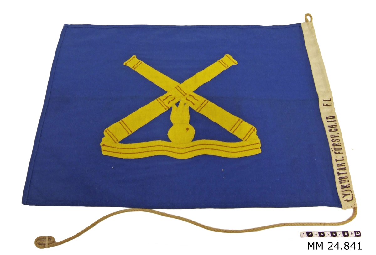 Rektangulär flagga i blått tyg. Påsytt finns Kustartilleriets emblem i gult med två korslagda kanoner. Under finns en eldfängd kanonkulla över vågor. På liket finns märkningen "Kustart.försv.CH I.D.". Vitt lik med snöre trätt igenom. Överst på liket är snöret i en ögla. Flaggan tillkom museet i en ram med lapp: 1992-09-29 "Denna flagga har tillhört batt TÖ och har enligt uppgift aldrig varit hissad. Om detta beror på att flaggstång saknats eller att försvarschefen aldrig har besökt förbandet framgår ej av historien. Flaggan överlämnad 1992 -09-29 på Kungsholmsfort  till Marinens officershögskola. Kn Sune Persberg."
