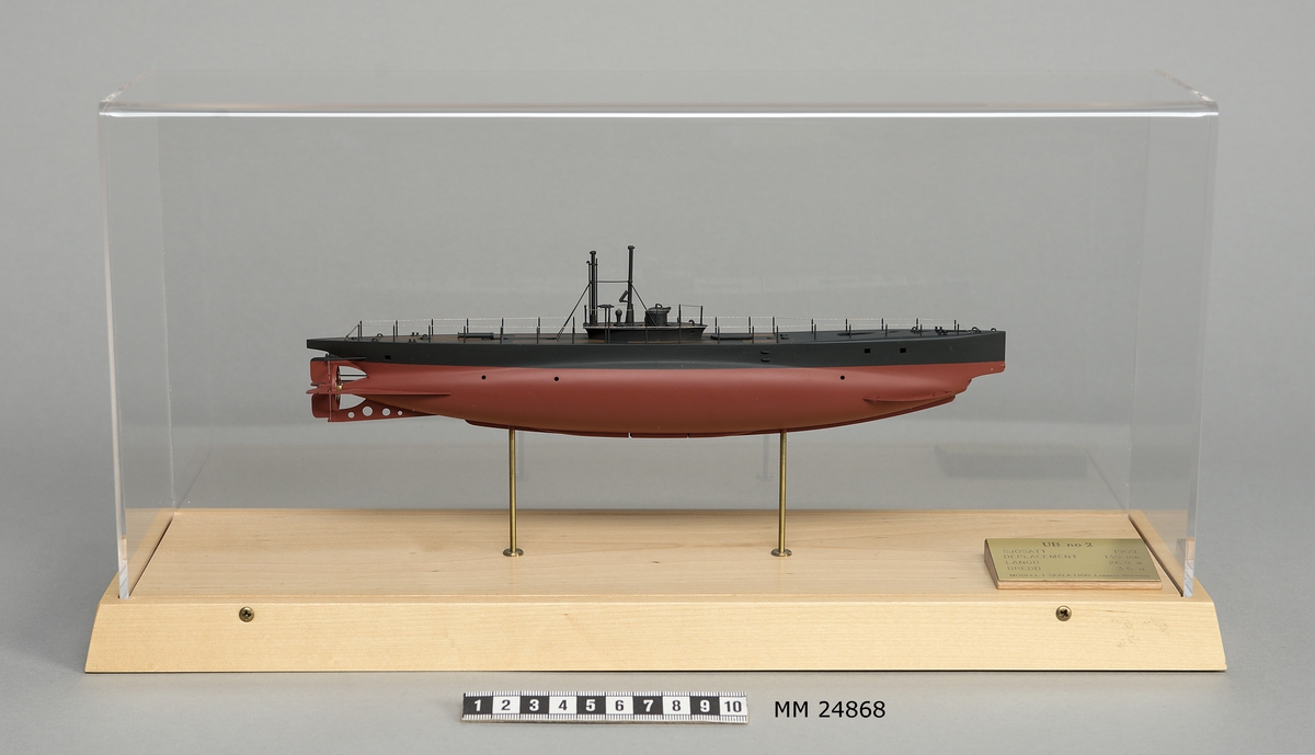 Ubåtsmodell UB No 2 i monter. Modell av alträ med detaljer av mässing, målad med cellulosafärg. Rött och svart skrov. Monter av plexiglas på träplatta. Mässingsbricka i montern med uppgifter om modellen.