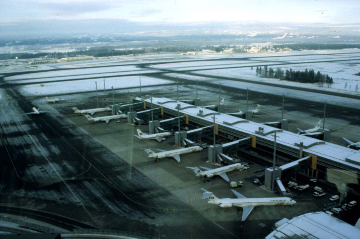 Lufthavn, litt av terminalen med flere fly, litt av rullebanen i bakgrunnen.