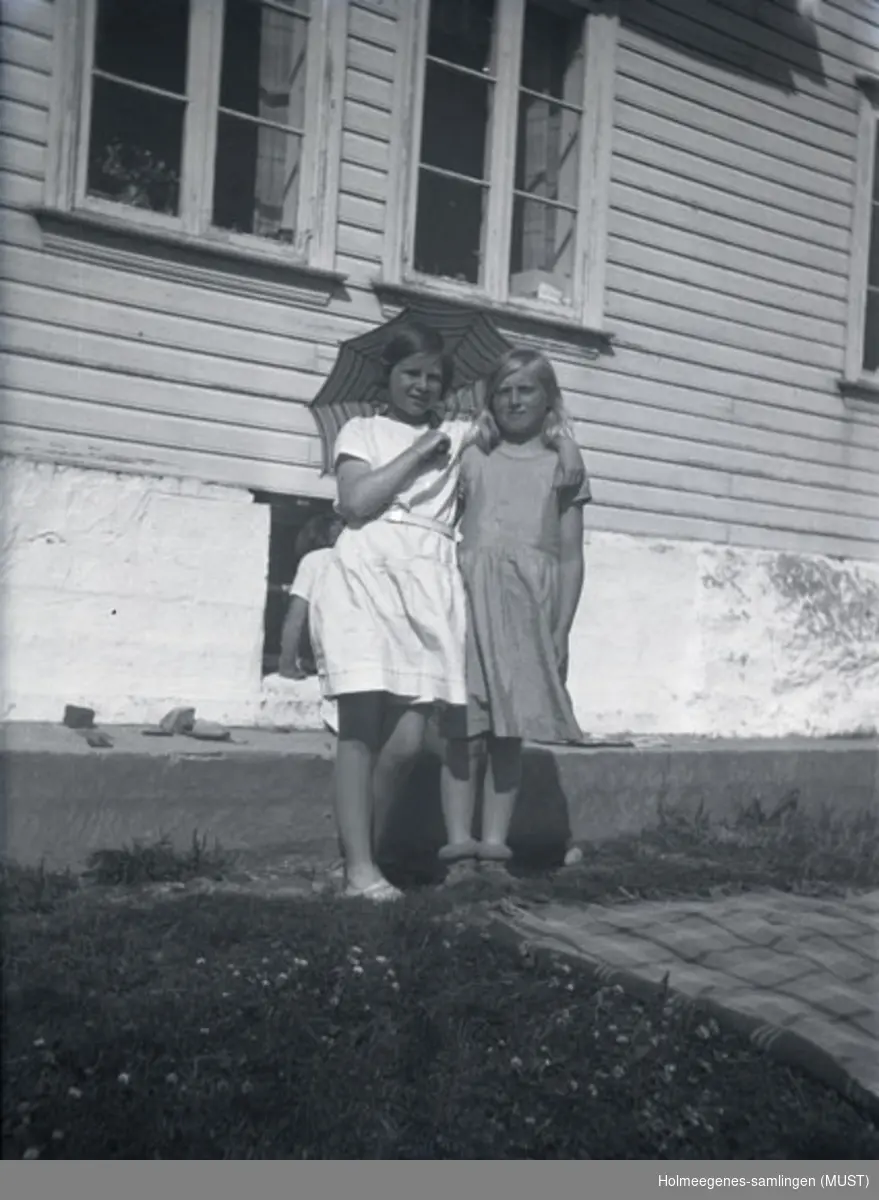 To jenter i sommerkjoler og med parasoll foran et bolighus. Til høyre for dem ligger det et pledd på gresset. Antatt samme sted og tidspunkt på ST.K.HE 2010-011-0096 til -0100.