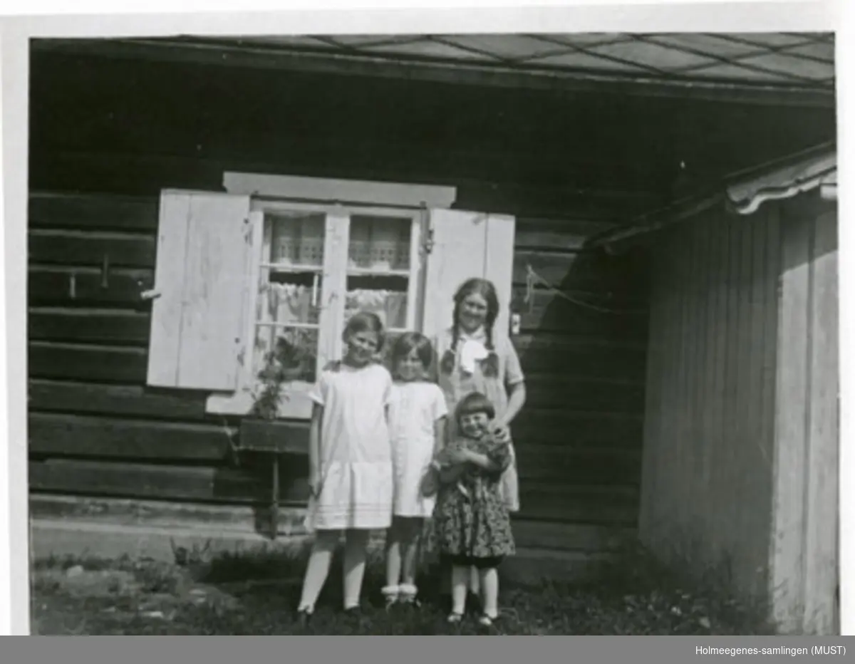 Fire jenter står ved en husvegg. Rett bak dem et vindu med skoddene åpne. Den minste jenta holder en katt i armene. Se også ST.K.HE 2007-011-0111 til -0126.