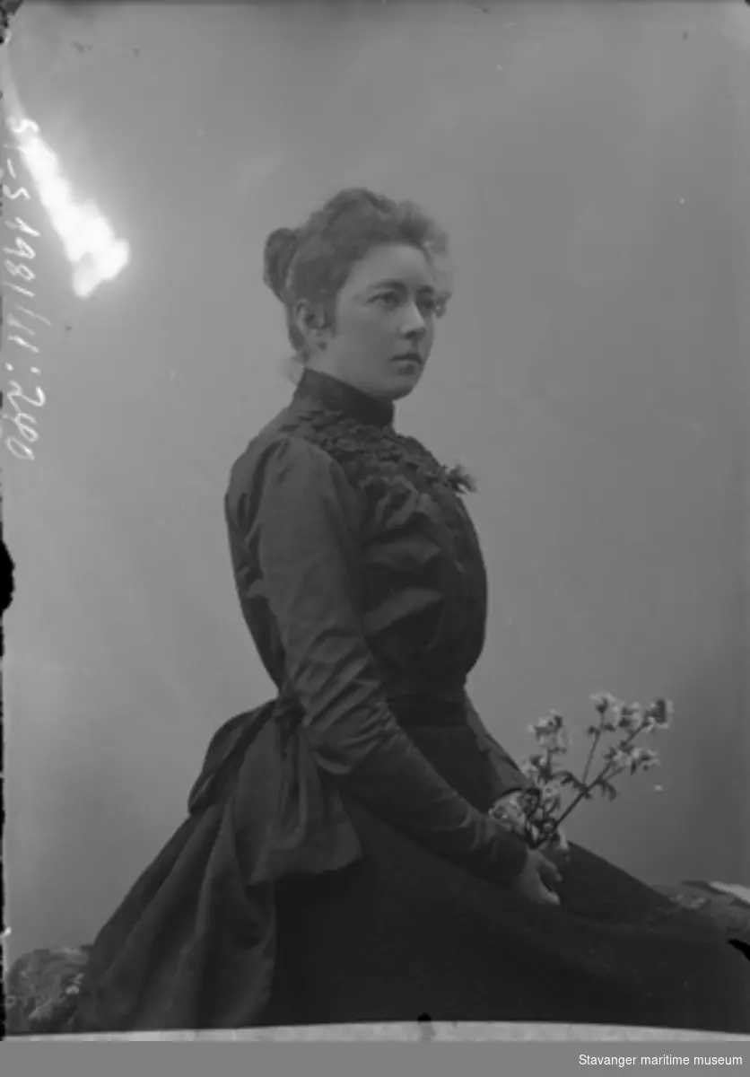 Studioportrett av ung kvinne i sort silkekjole. Sittende i profil, halvfigur. Nøytral bakgrunn.