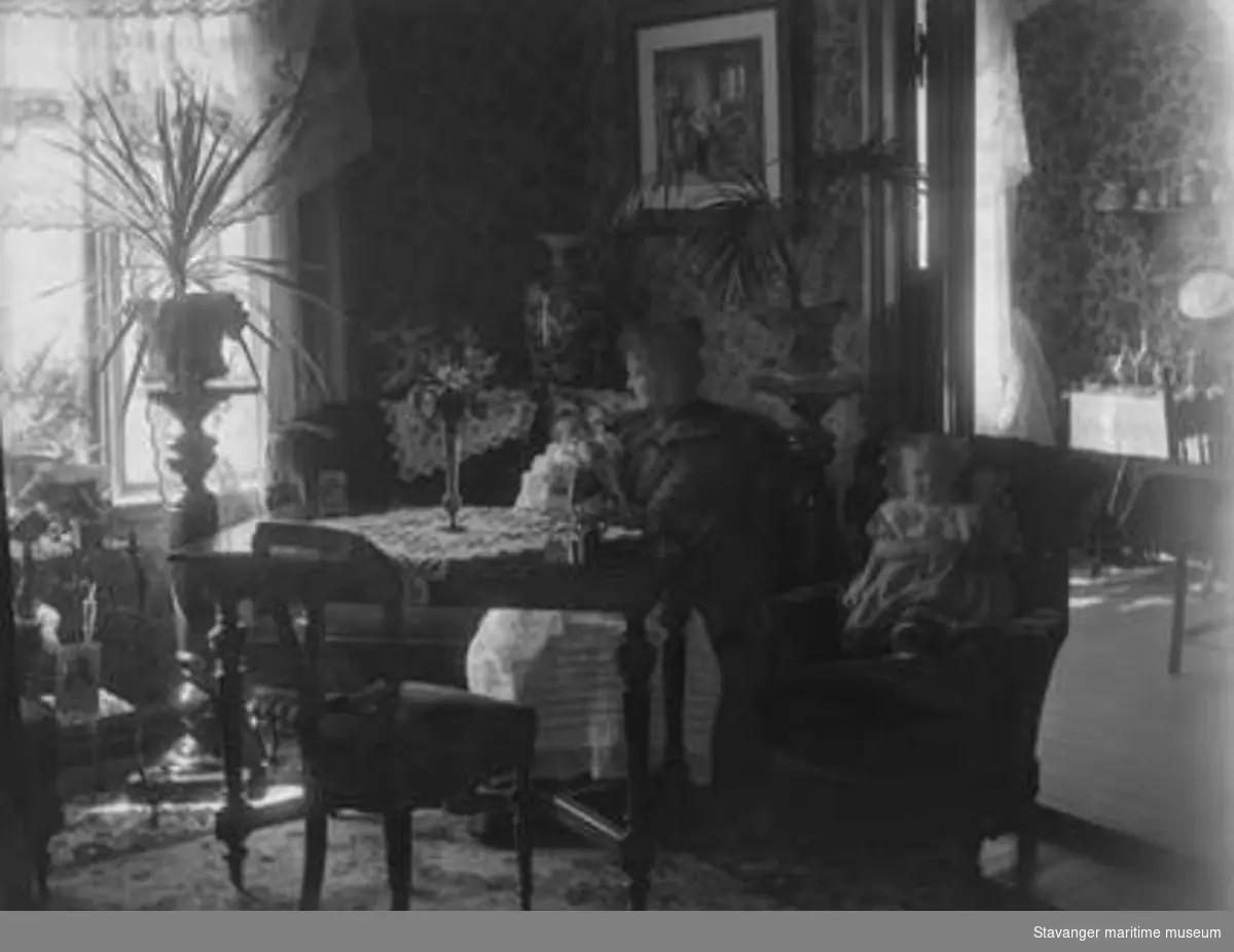 Borgerlig interiør. Kvinne med spedbarn i dåpskjole sitter i sofa. På bordet står fotografier oppstilt. Barn på ca 2 år sitter på stol.
