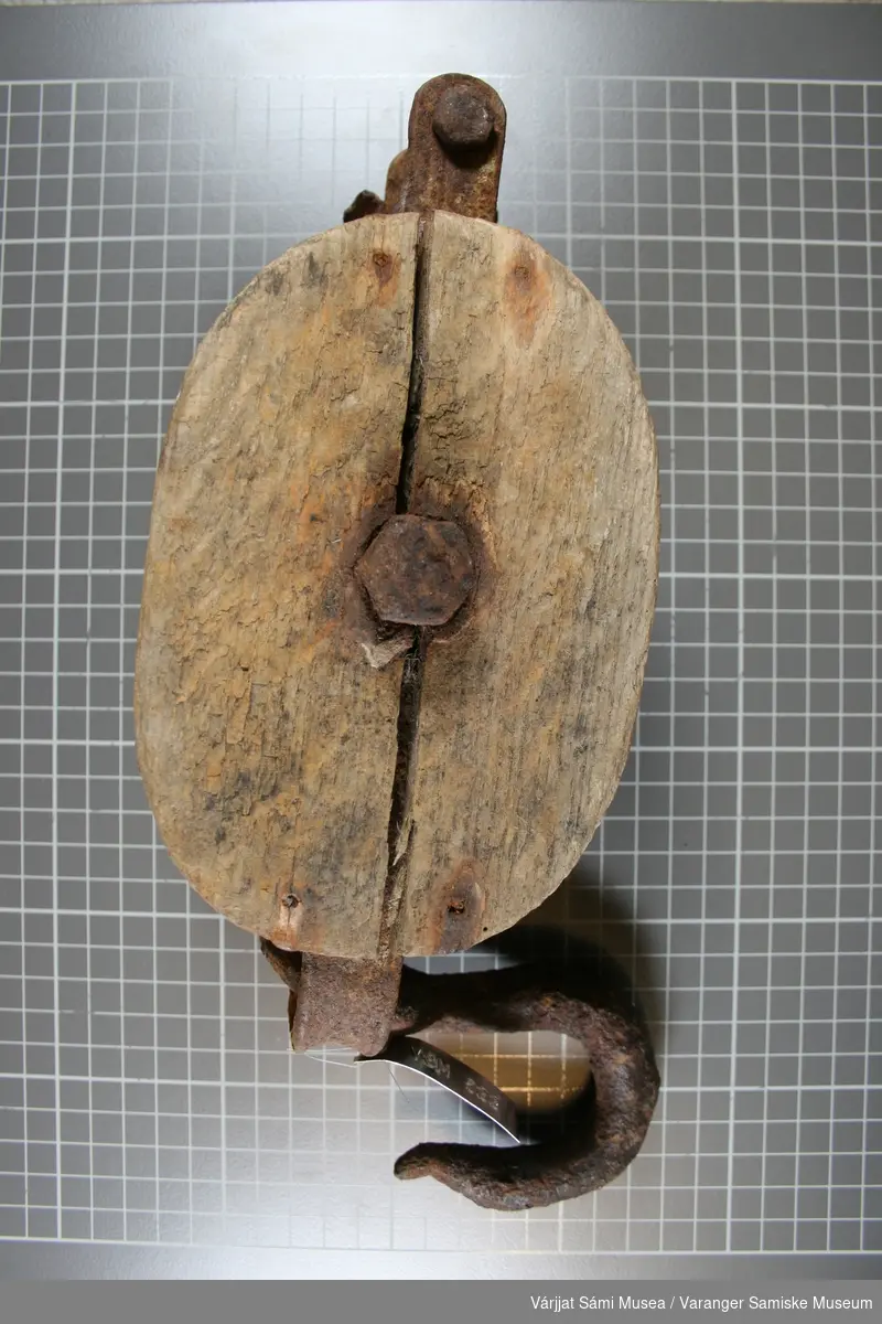 Talje av tre og jern. Taljen (den runde skiva hvor tauet går rundt),  kroken og opphenget er av jern.
