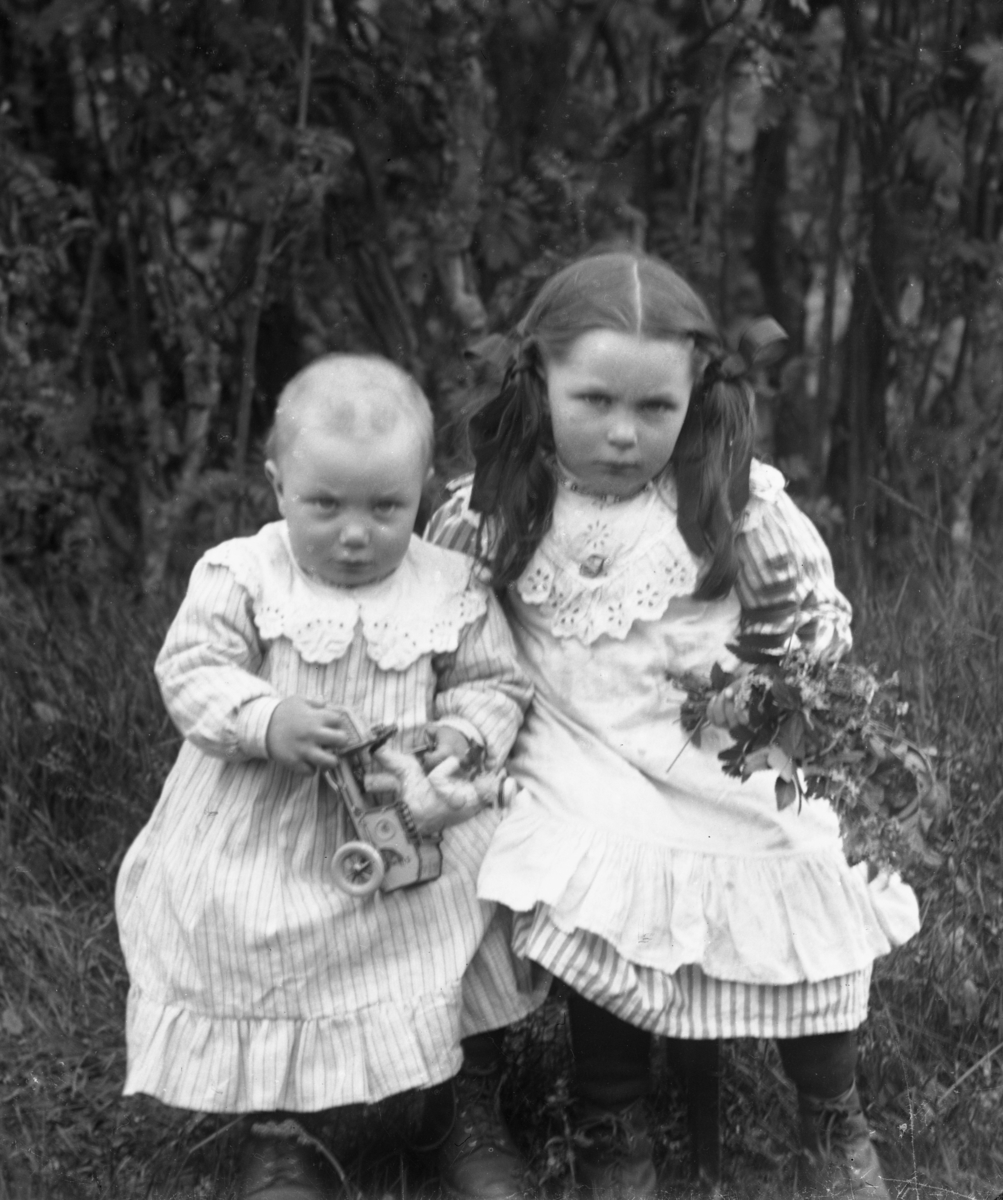 Liten jente med musefletter, holder blomsterbukett, samt mindre barn med lekebil, begge kledd i hvite kjoler. Gress og busker i bakgrunnen