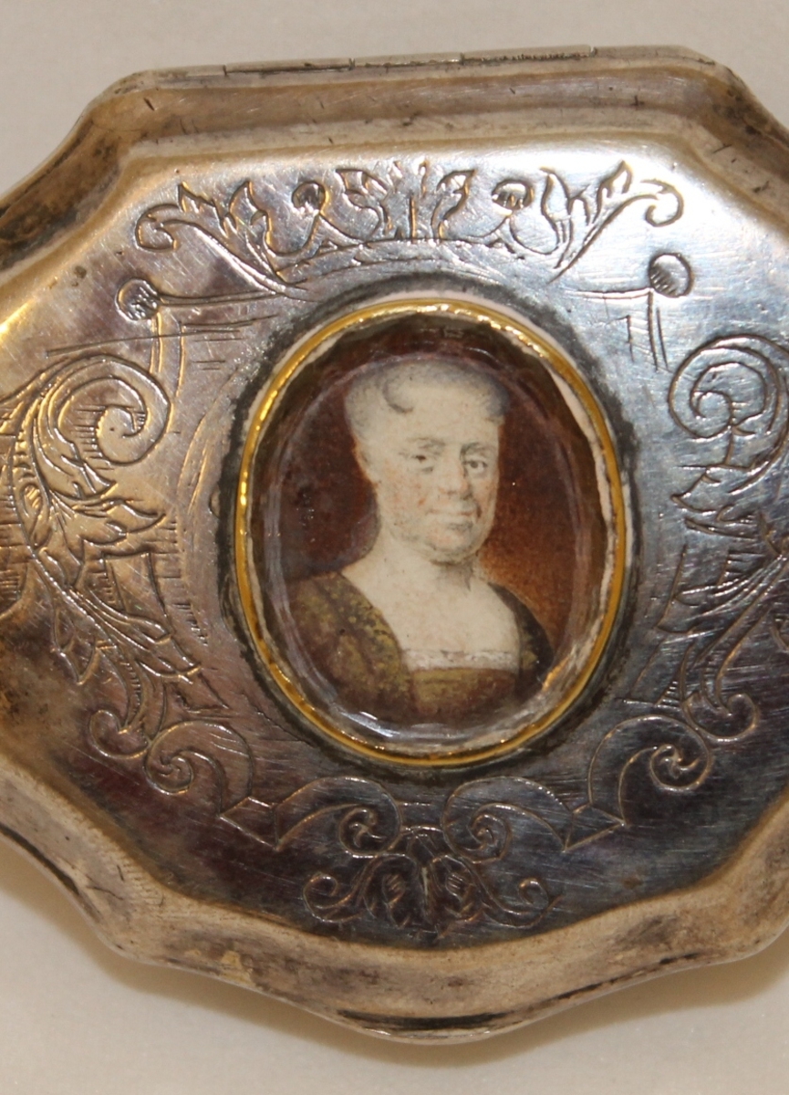 Kvinne 3/3 en face, grå, flat frisyre med pannelokk, firskåren utringning. 
Dekket av ovalt glass med fasettslepen kant og gull-kant.(Hopstock: Ant. Madame Anna Adeler (død 1729)