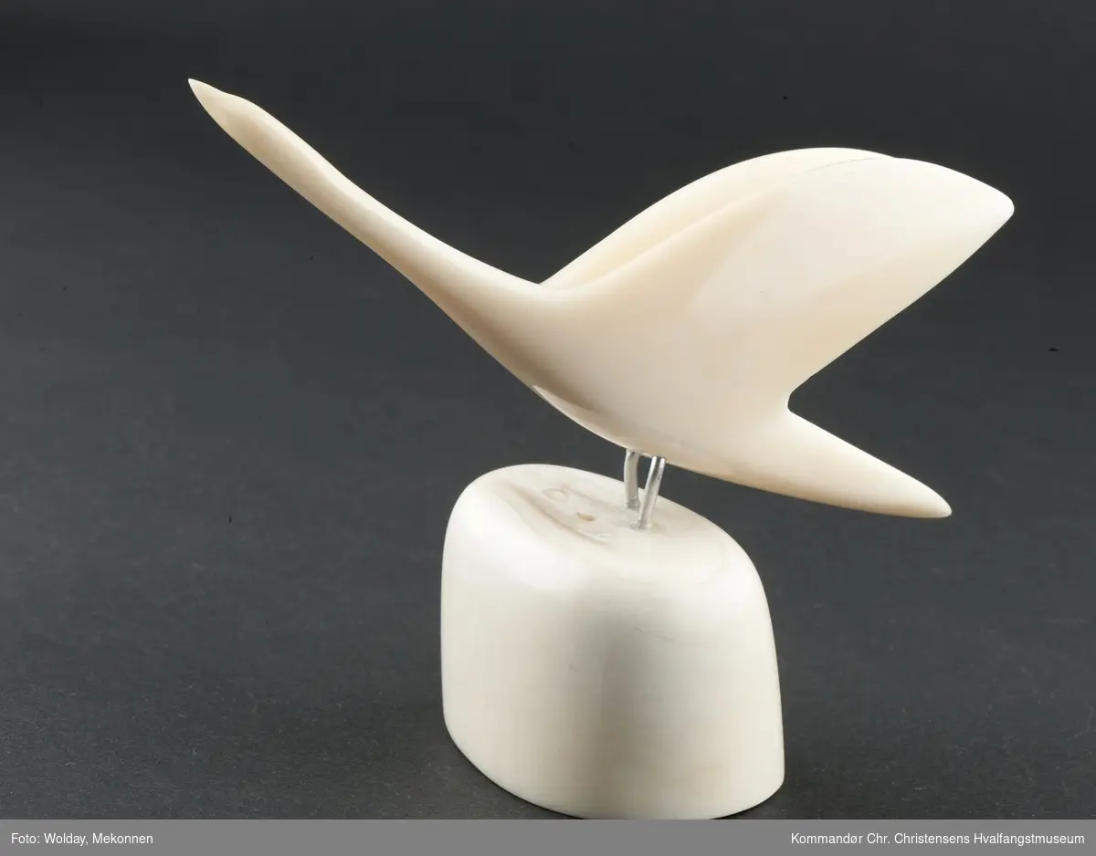 Stilisert fugl. Framstilt av hvaltann. Utskåret, slipt, polert. Figur festet til sokkel med metall- stenger