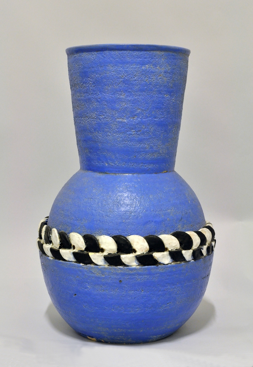 Stor vase av "keramikk". Tilnærmet kuleform med høy, vid hals. På midten et bånd av sirkulære plater som er kjedet i hverandre, med vekselvis sort og hvit glasur. Vasen har en ru overflate og er malt med blå farge. Form: Nora Gulbrandsen.