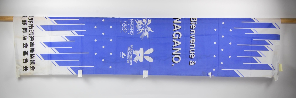 Blått, hvitt og sort avlangt banner med logo for de olympiske leker i Nagano i 1998 og Paralympics i Nagano i 1998. Banneret har en del slitasje og flekker. 