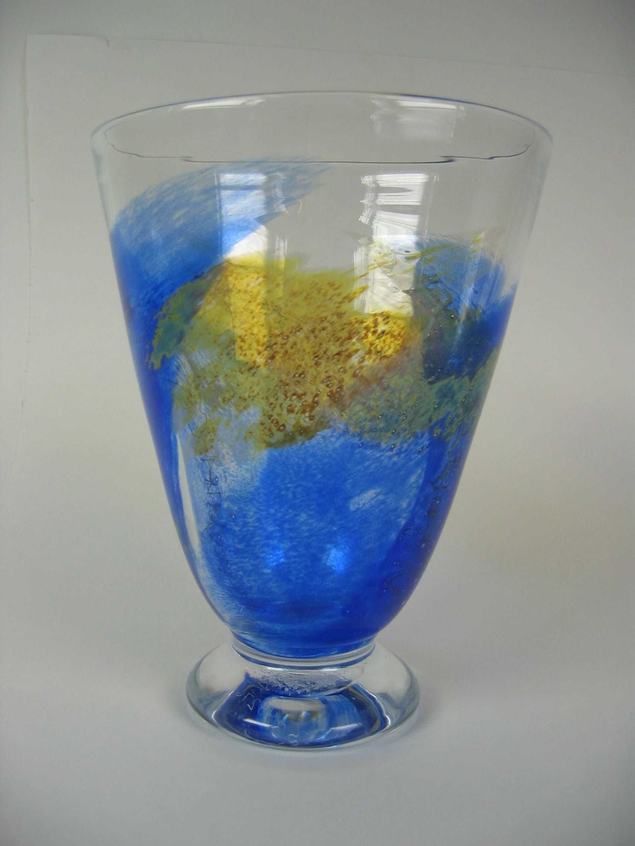 Lett flatklemt konisk formet vase på en liten fotplate. Blåst i to deler. Hovedfargen er klart glass, med noe blått og brunspettet.