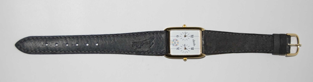 Gull- og sølvfarget armbåndsur med sort reim og hvit urskive. På urskiven er det et emblem med motiv av de olympiske ringene og en stilisert sol. Det følger med et grått etui.