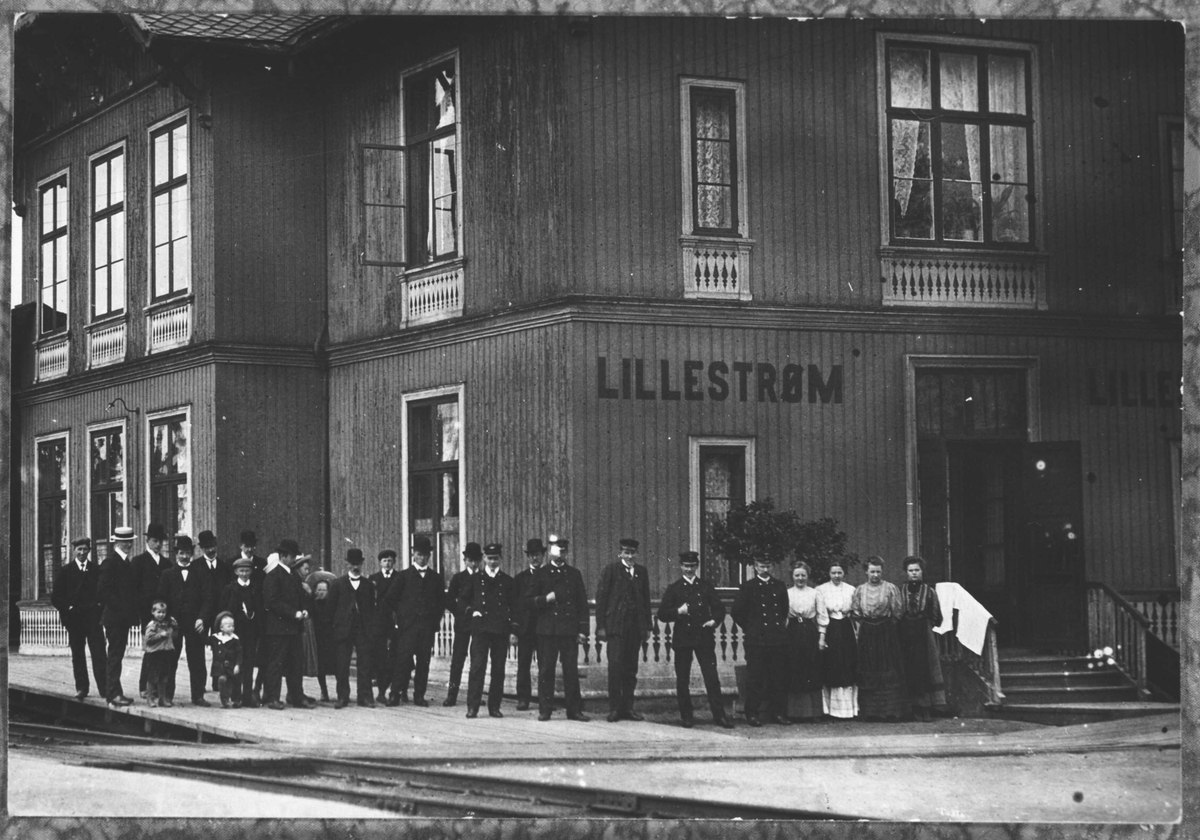 Lillestrøm stasjon. Den første stasjonsbygningen på Lillestrømsiden.
Jernbanepersonale i uniform, samt en stor flokk sivile. Fire damer (hotellbetjening?) de øvrige er herrer i dress, og tre fire små barn.