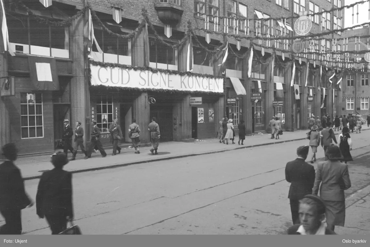 Folkevandring i en pyntet Rådhusgate 7. juni 1945 i anledning Kongens hjemkomst. Banner med teksten "GUD SIGNE KONGEN". Girlandere.
