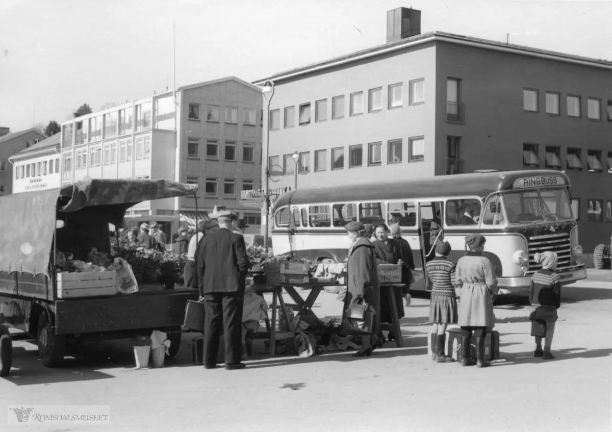 Torgsalg i Molde..Hjelset Auto startet Ringbuss i Molde i 1953, de første sjåførene var Bjørn Hammerø og Knut Stranden. .Det var i mange år bare to ruter, Østre og Vestre ring. Det var avgang hver time på hver rute. .Mellom turene var et pause ved holdeplassen ved Torget. Bussen har dermed kommet vestfra og venter på avgang østover (Kviltorp) da bildet ble tatt.