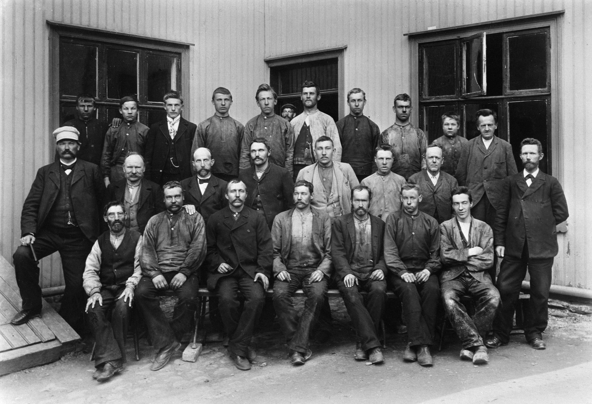 Gruppe 25 arbeidere, Lunds Vognfabrikk i Hamar, håndverkere. T. v. Andreas Lund med skyggelue, mannen midt i bildet med lys dress og klokkelenke er Ole Peder Holter. 