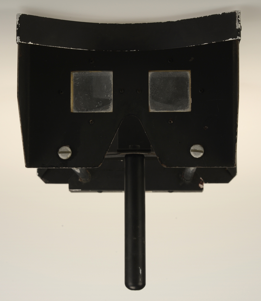 Stereoskopapparat med to metallplatersom er forbundet med to runde stenger. Kvadratiske glasslinser i metallplate nærmest øyet og plate for innsetting av stereoskopikort. Denne er regulerbar i avstand fra linsedelen. Håndtak nærmest linsedelen. Med stereoskopiske kort.